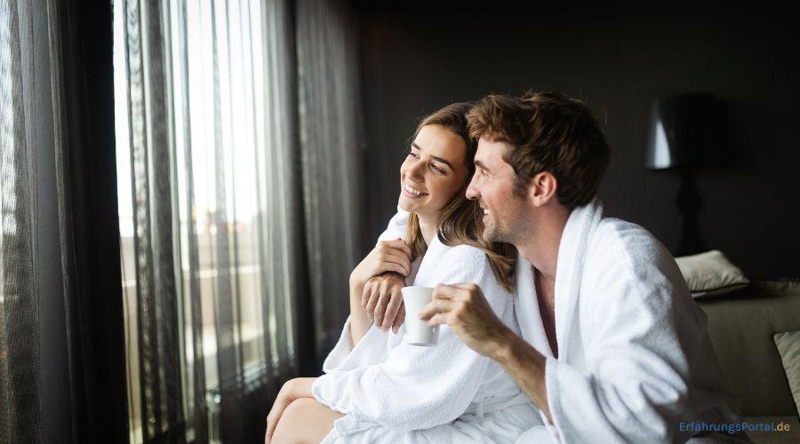 Ein Paar sitzt in Bademänteln auf einem Bett und schauen mit einem Lächeln im Gesicht aus dem Fenster. Der Mann hat die Hand um die Frau gelegt und in der anderen Hand hält er eine Tasse