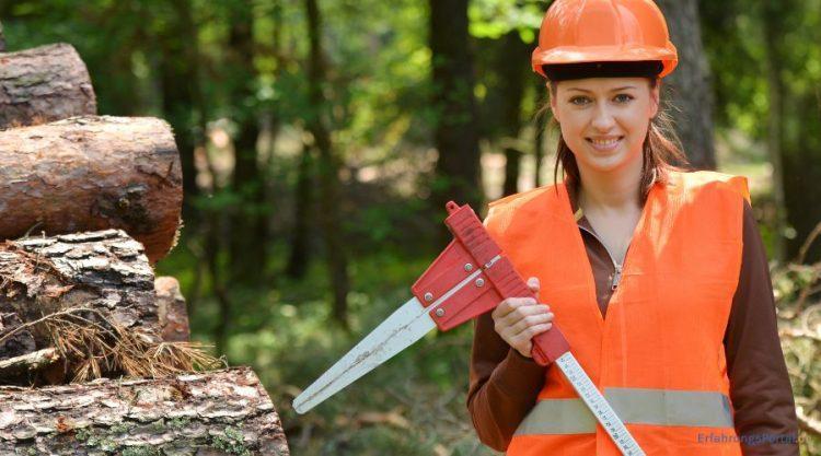 Frau mit Warnweste und Sicherheitshelm steht neben gefällten Baumen die auf einen Haufen gelegt worden sind