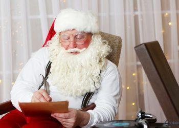 Der Weihnachtsmann sitzt auf einem Sessel und liest einen Brief. Er hält eine Feder in der Hand. Neben ihm steht ein Plattenspieler