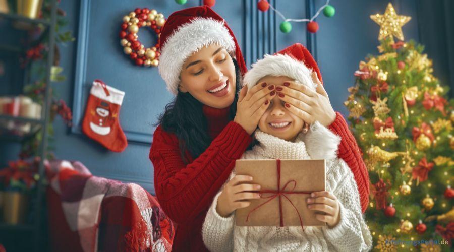 Mutter mit Kind unter dem Weihnachtsbaum. Das Kind hält ein verpacktes Geschenk in der Hand, während die Mutter ihm die Augen zu hält. Beide lachen und tragen eine Weihnachtsmütze. Im Hintergrund steht ein geschmückter Weihnachtsbaum