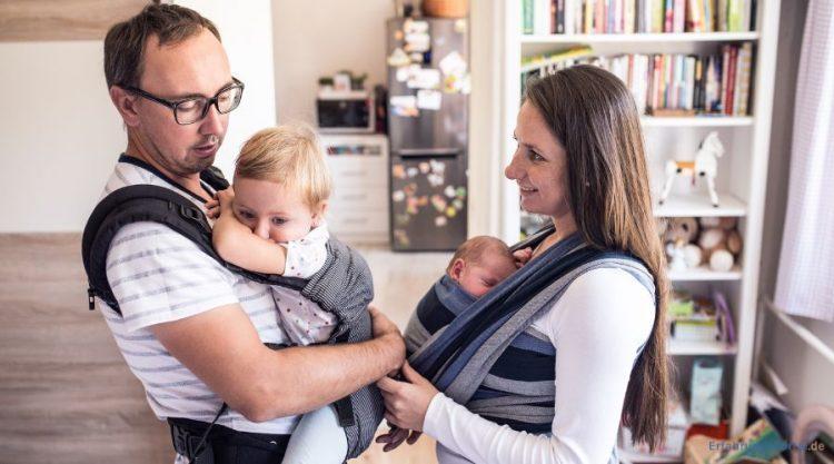 Eltern tragen ihre zwei Kinder in einem Tragetuch und in einer Trage