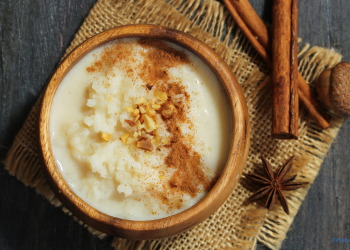 Milchreis mit Zimt wurde in einer Holzschüssel angerichtet. Neben der Schüssel liegen Nelken und Zimtrollen